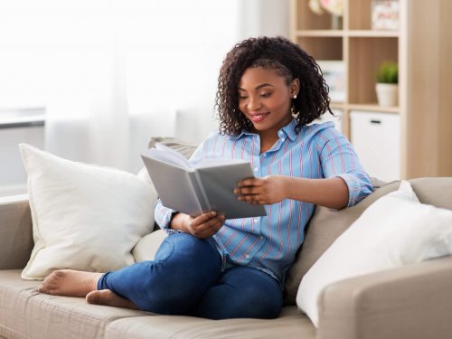 leisure, literature and people concept - smiling african american woman reading book at home